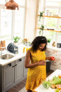 A pregnant women holding  a tomato and preparing food in a well-lit kitchen